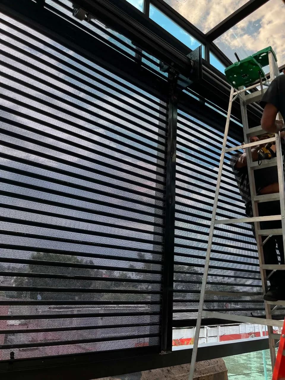 Instalador en escalera montando una cortina perforada negra en una terraza con techo de cristal